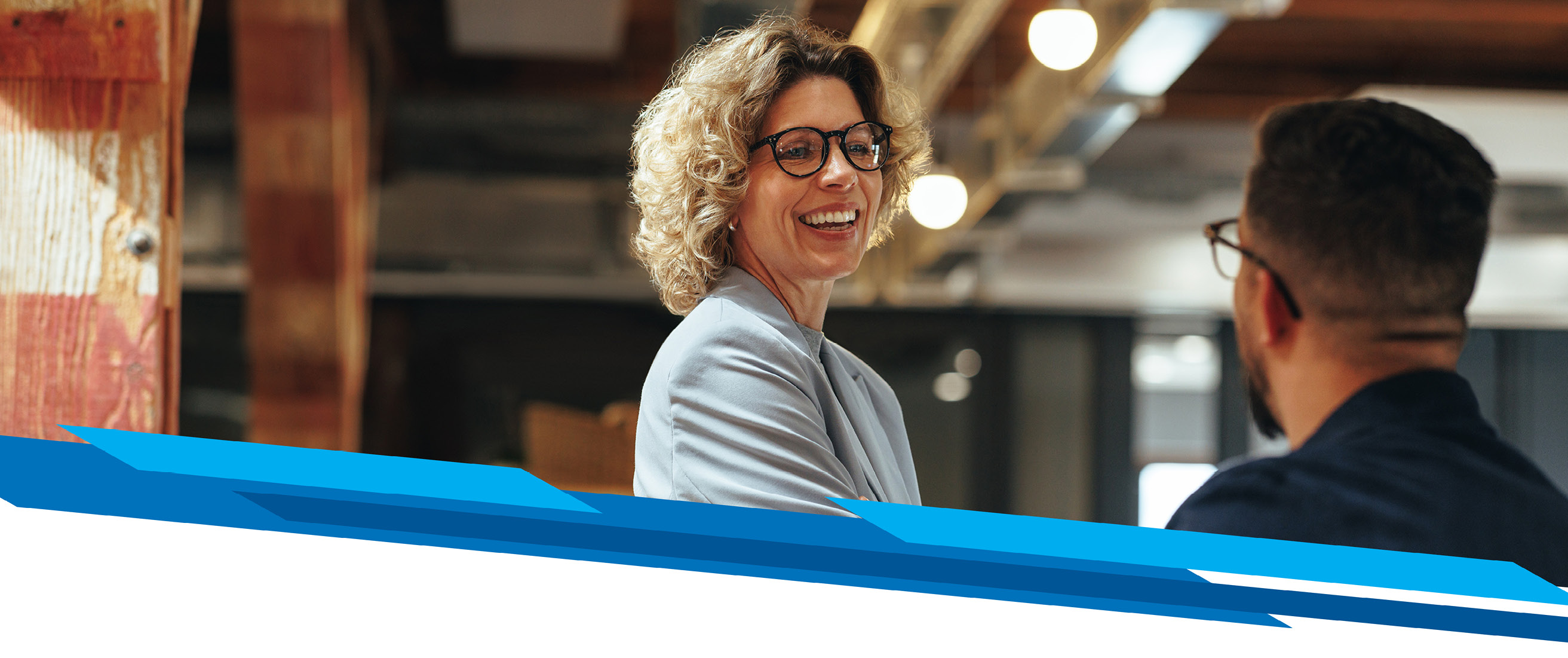 A professional woman with blonde curly hair and glasses is smiling at her colleague, expressing approval of a successful business discussion. 